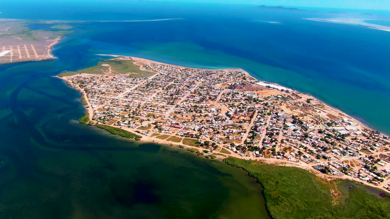 A high aerial view over San Carlos, Baja California Sur shows a compact town on a narrow peninsula, shallow green water in front, deeper blue water beyond, and no visible people.