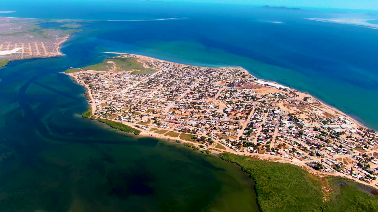 This aerial frame shows San Carlos, Baja California Sur from above, with the town angled across the peninsula, green vegetation along the lower shoreline, blue water to the right, and no visible people.