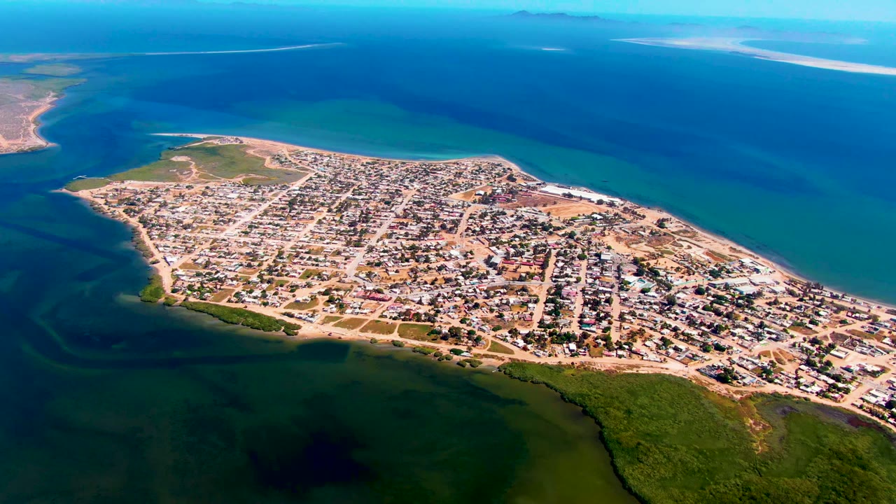 High aerial view over San Carlos, Baja California Sur, showing a gridded coastal town on a sandy peninsula with green shoreline vegetation in the foreground and no visible people.
