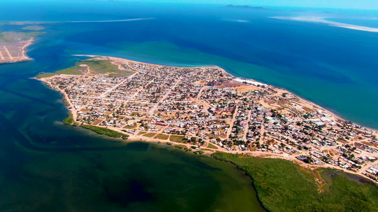 Wide drone view of San Carlos, Baja California Sur, showing the peninsula settlement surrounded by deep blue and turquoise water, with pale sandbars near the distant horizon and no visible people.