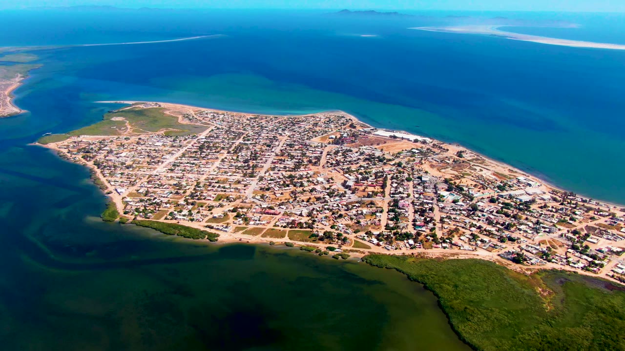 Aerial view over San Carlos with the peninsula town centered, calm blue water stretching to the horizon, darker shallows in the foreground, and no visible people.
