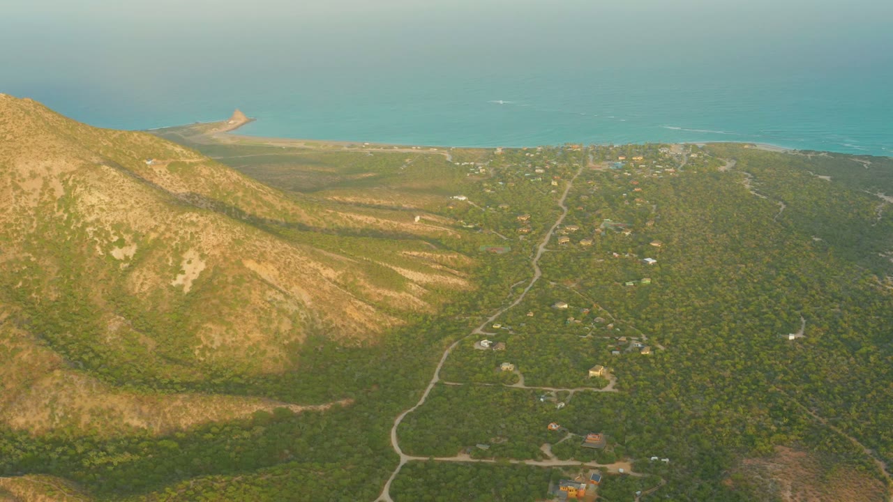 Aerial view over Cabo Pulmo, Baja California Sur, with a tan mountain slope on the left, green forest and winding roads below, blue ocean at the horizon, and no visible people.