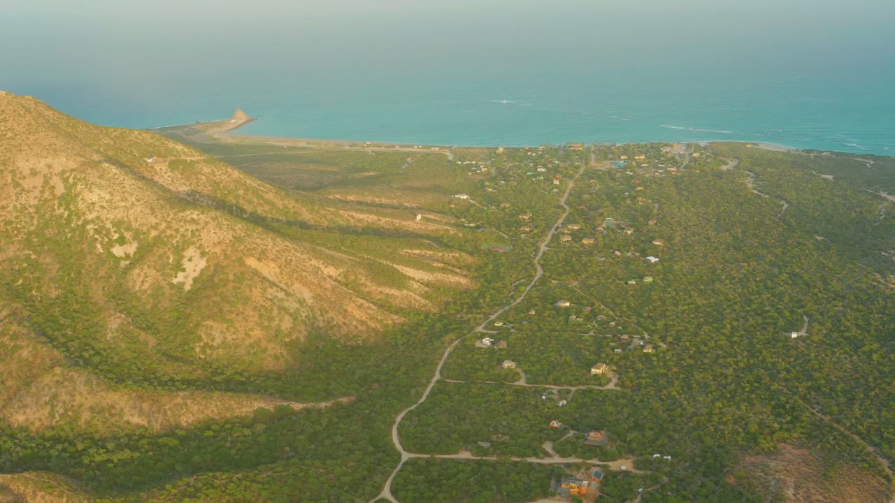 Wide aerial landscape with mountain slope, forest roads, scattered buildings, and ocean beyond.
