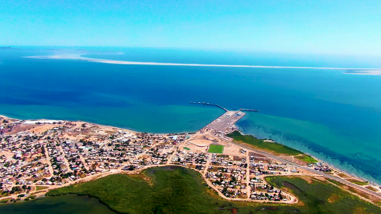 Opening aerial frame over San Carlos, Baja California Sur, with houses and roads below, a pier extending into clear blue water, a long pale shoreline on the horizon, and no visible people.