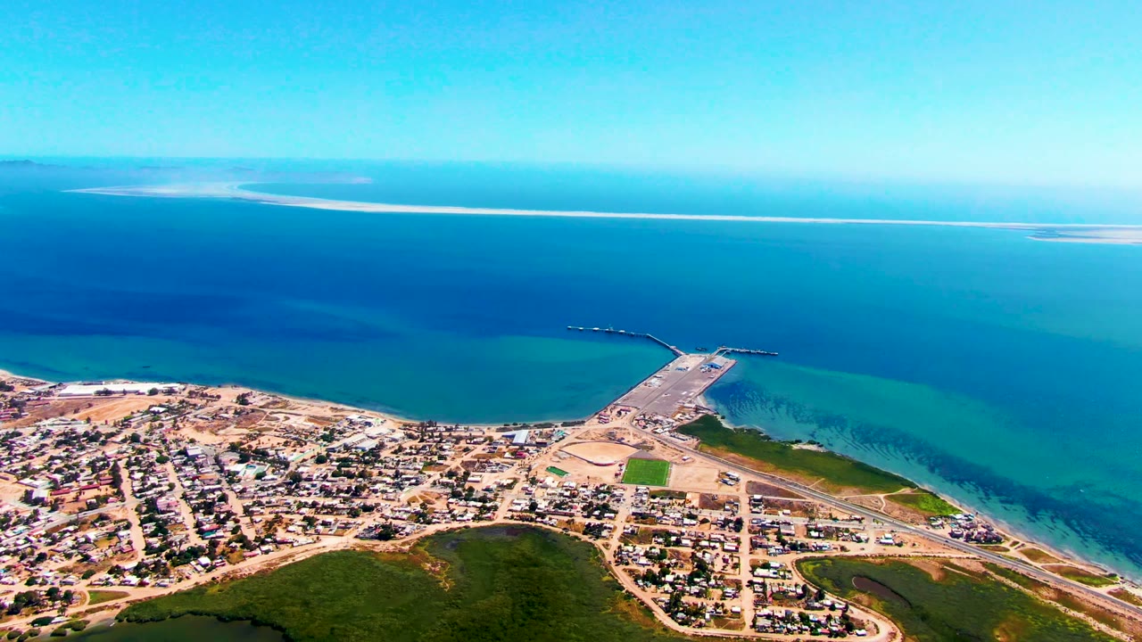 Aerial view in San Carlos with residential blocks at the bottom, the pier angled toward the ocean, shallow green-blue water near shore, darker water beyond, and no visible people.