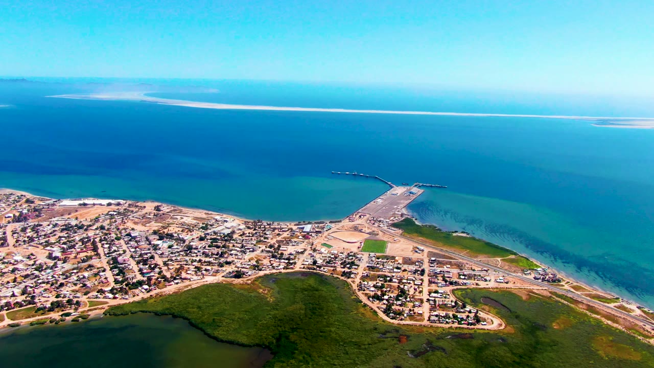 Opening aerial frame over San Carlos, Baja California Sur, showing town blocks, sandy shore, a pier or breakwater, green coastal vegetation, blue ocean, distant sandbars, and no visible people.