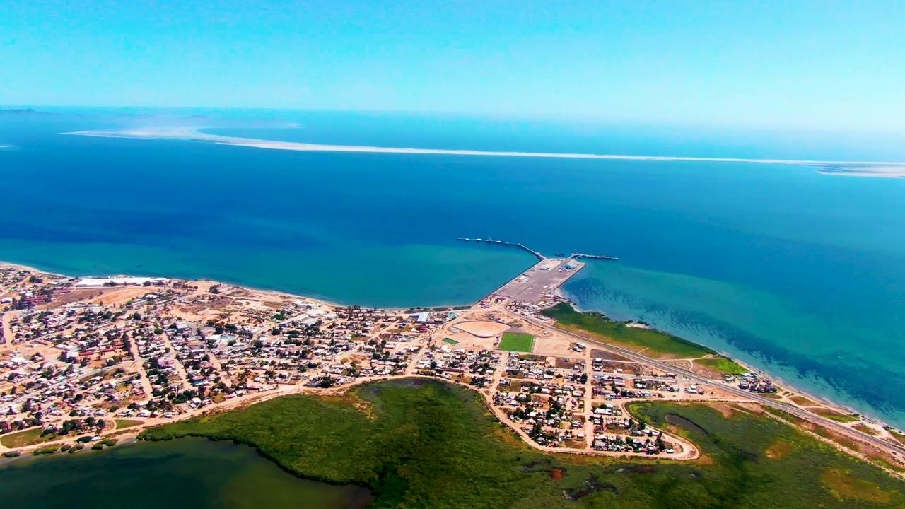 Wide drone view of shoreline town, green vegetation, pier, and calm blue water.