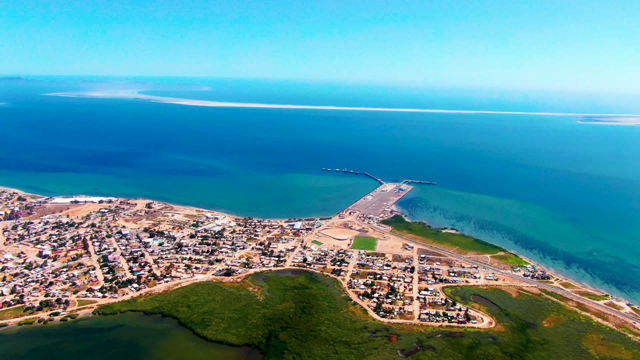 At 0:02, the San Carlos shoreline remains in a broad aerial composition with clustered buildings, pale beach, turquoise shallows, darker offshore water, distant sandbars, and no visible people.