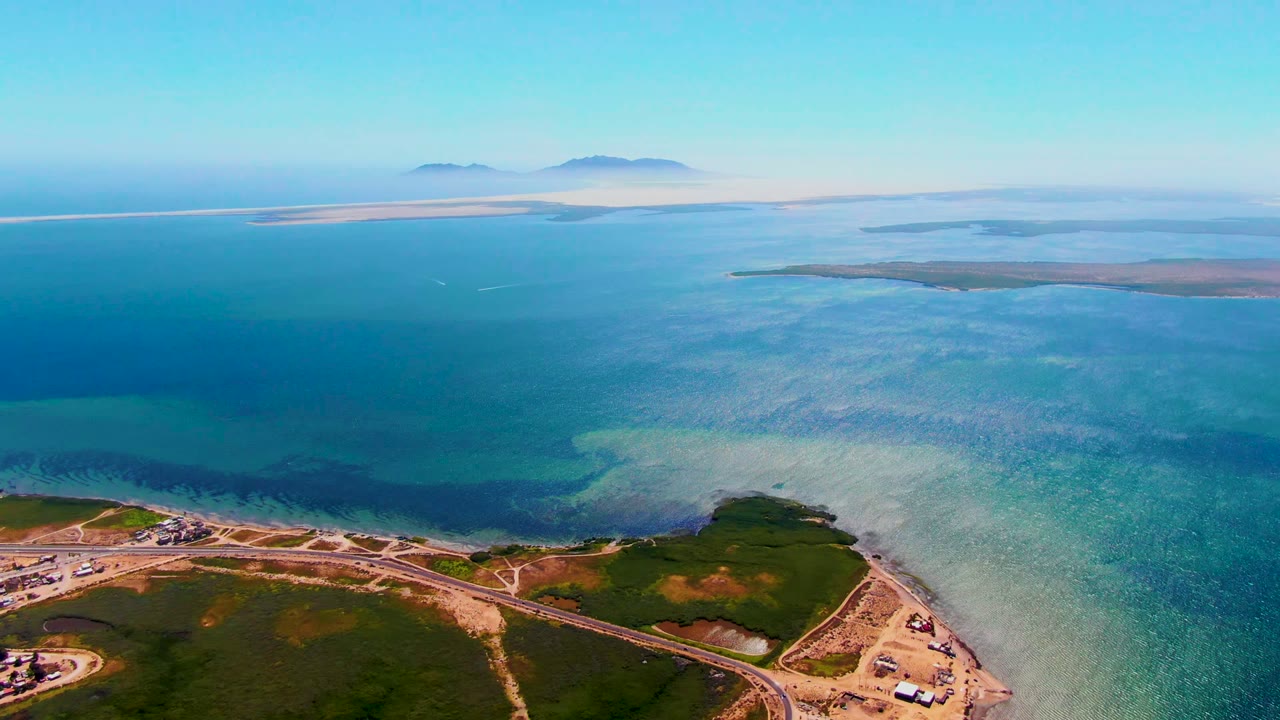Opening aerial frame over San Carlos, Baja California Sur, with a curving shoreline road, green land, turquoise shallows, distant islands, and no visible people.