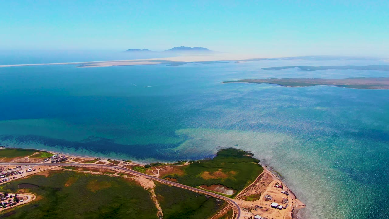 Mid-clip aerial frame of San Carlos coastline with the road and small developed areas at the bottom, clear blue water across the center, distant islands, and no visible people.