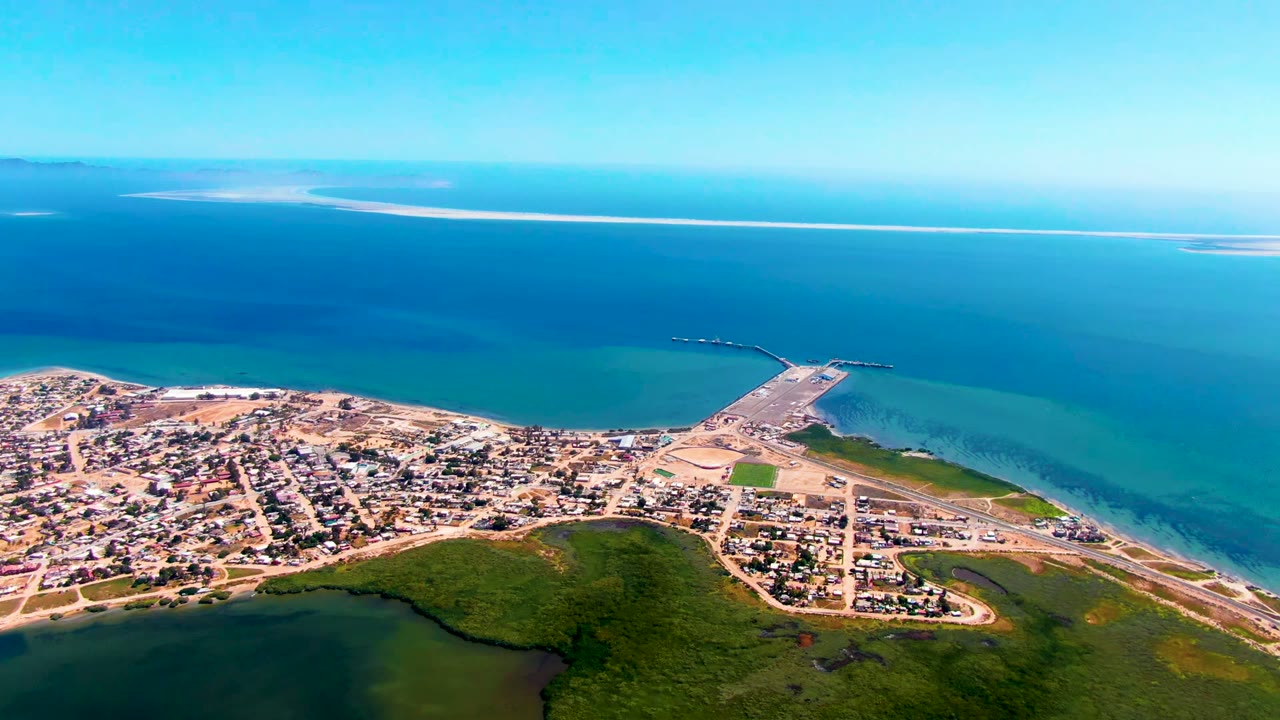 Wide aerial view over San Carlos, Baja California Sur, showing residential blocks, green foreground land, a harbor extending into blue water, distant sandbar, clear sky, and no visible people.