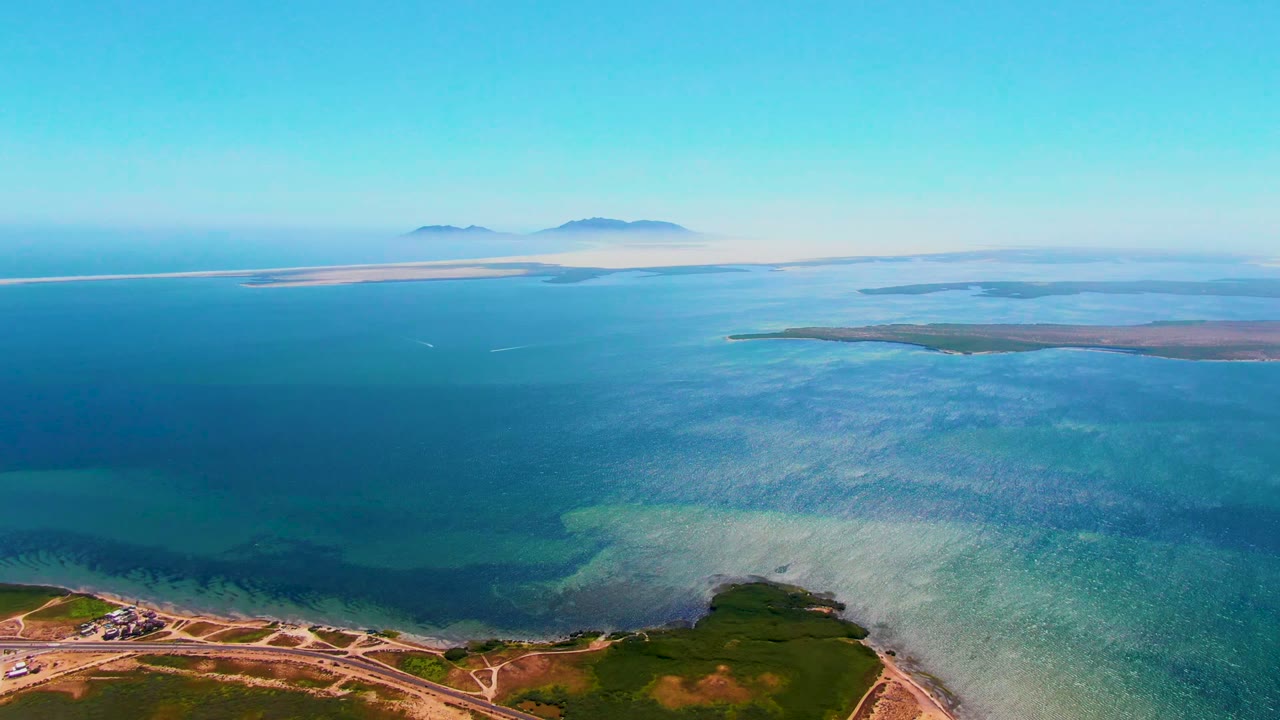High aerial frame over San Carlos, Baja California Sur showing a coastal road and green land below, turquoise shallows, deeper blue ocean, distant islands on the horizon, and no visible people.