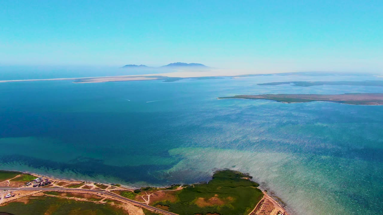Wide aerial coastline with road, shallow turquoise water, deep ocean, and hazy islands.