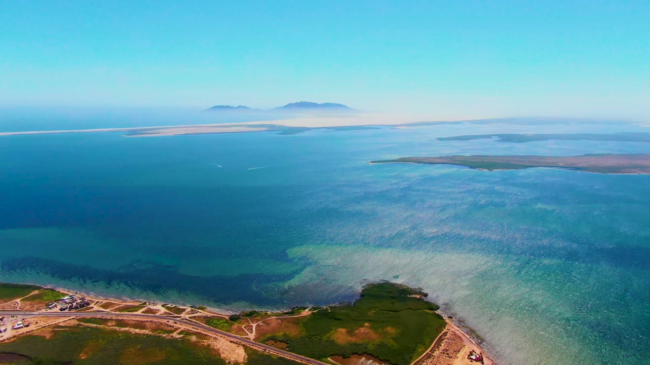 High drone frame over San Carlos, Baja California Sur showing more of the coastal road network at the bottom, green shoreline, layered turquoise and blue water, distant islands, and no visible people.