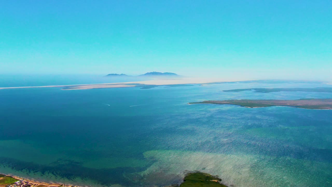 Aerial view over San Carlos, Baja California Sur, showing blue coastal water, hazy distant islands, a thin shoreline at the bottom edge, and no visible people.