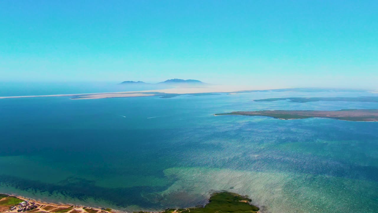 High coastal view with turquoise shallows, open blue water, distant islands, and shoreline tracks below.