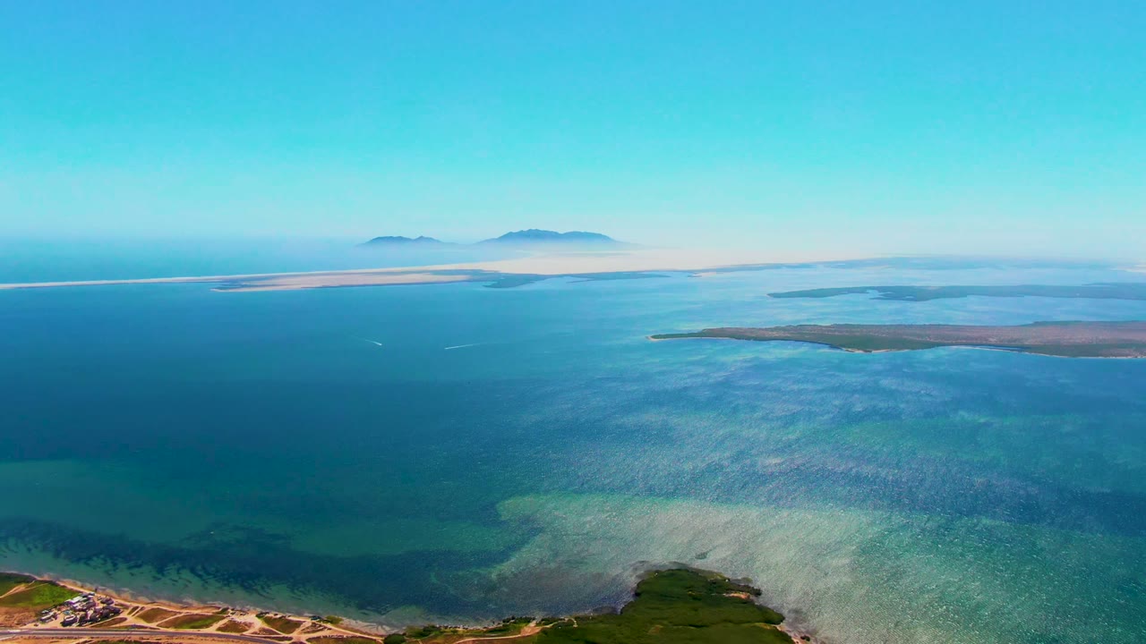 San Carlos, Baja California Sur aerial view with green foreground land at the lower right, layered turquoise and blue water, distant misty islands, and no visible people.