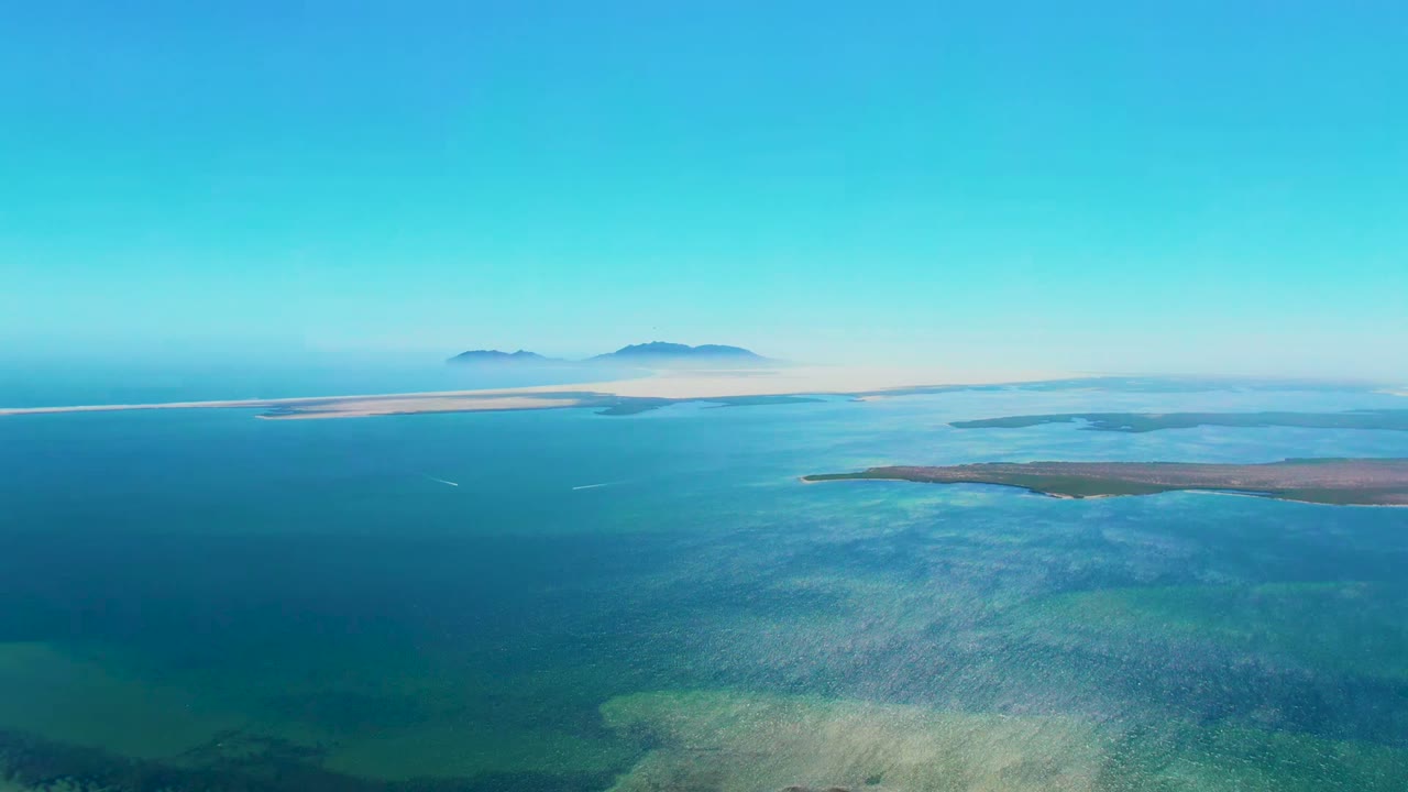 Opening aerial frame over San Carlos, Baja California Sur, with turquoise shallows in the foreground, darker blue water beyond, low land across the midground, distant islands on the horizon, and no visible people.