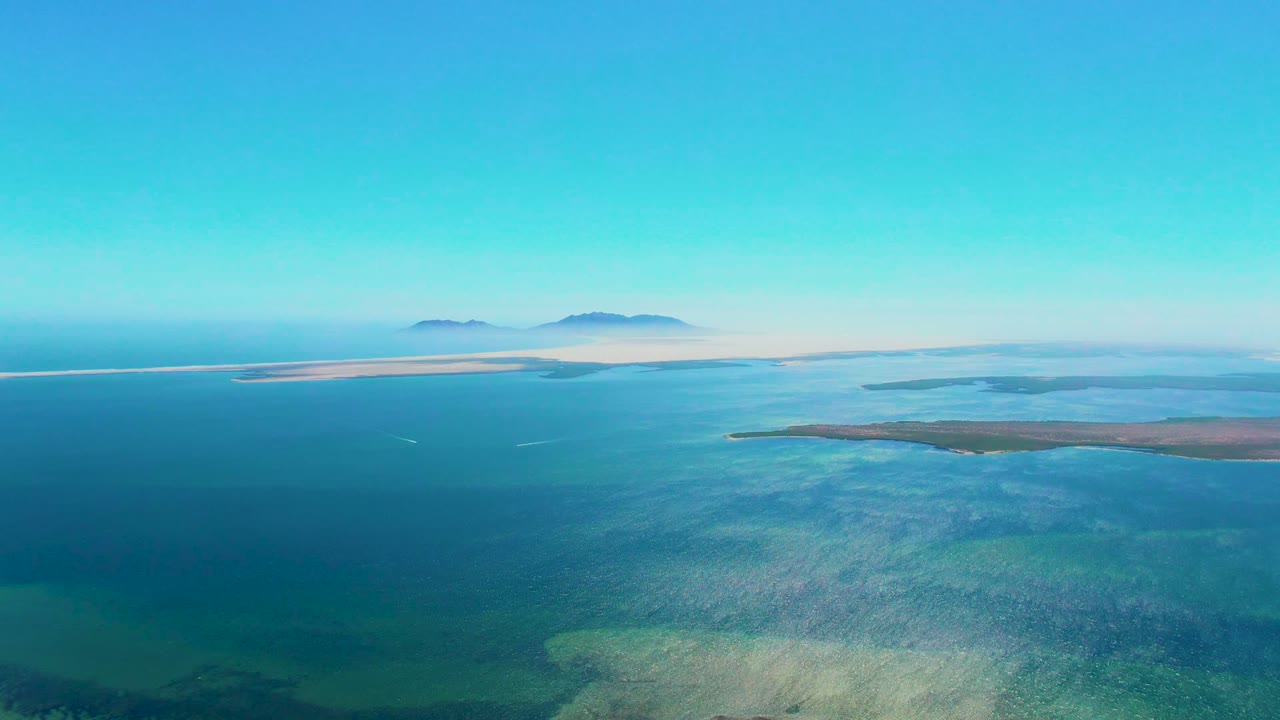 Wide aerial coastal view with visible seabed, low landmass, and hazy islands.