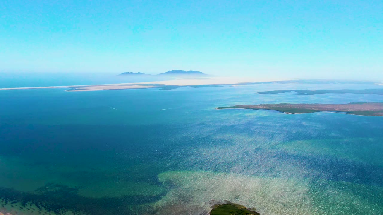 Opening aerial frame over San Carlos, Baja California Sur, with deep blue water in front, turquoise shallows near the lower edge, low islands across the middle distance, and no visible people.