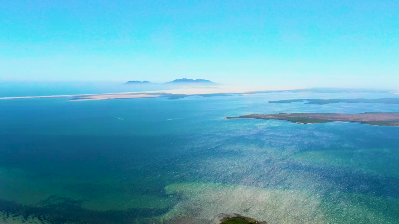 Wide aerial view over clear ocean with shallow turquoise patches and islands near the horizon.