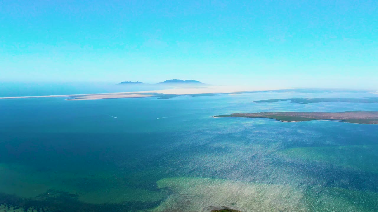 Mid-clip aerial frame over San Carlos, Baja California Sur, with a broad turquoise shelf in the foreground, deeper blue water beyond, low islands to the right, sandbars near the horizon, and no visible people.