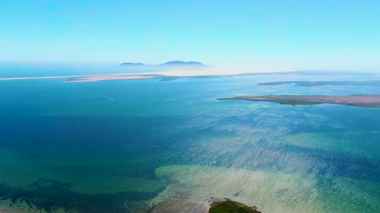 Aerial view over San Carlos, Baja California Sur, showing turquoise shallows in the foreground, darker blue water beyond, distant island silhouettes, and no visible people.