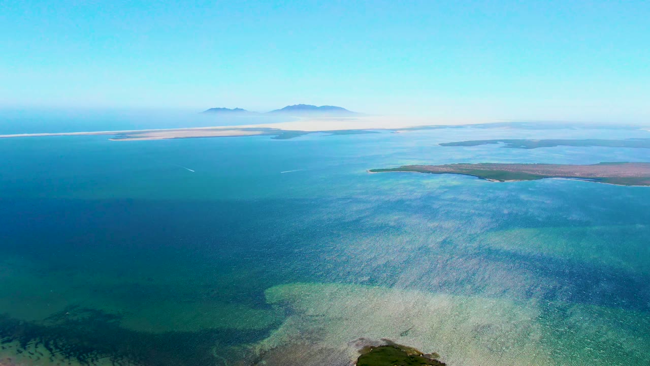 Wide aerial over shallow coastal water with islands on the horizon.