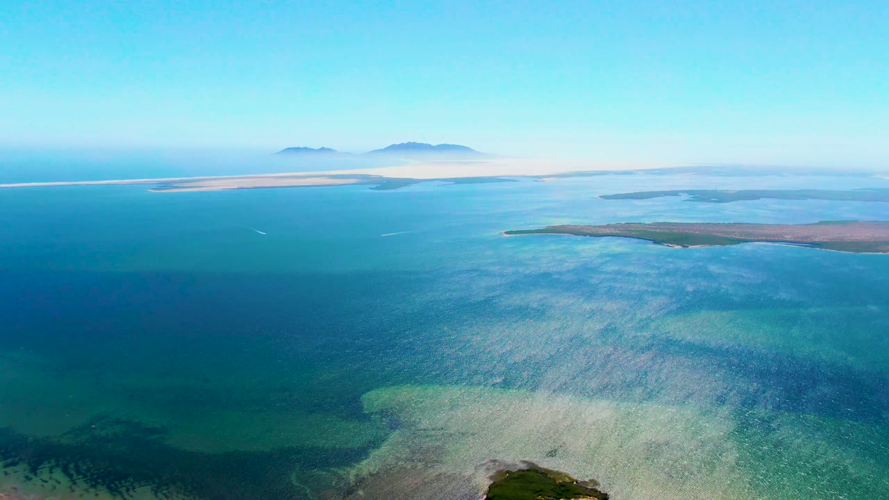 Aerial coastal view in San Carlos, Baja California Sur, showing a long pale sandbar near the horizon, layered blue water, a low landmass at right, and no visible people.