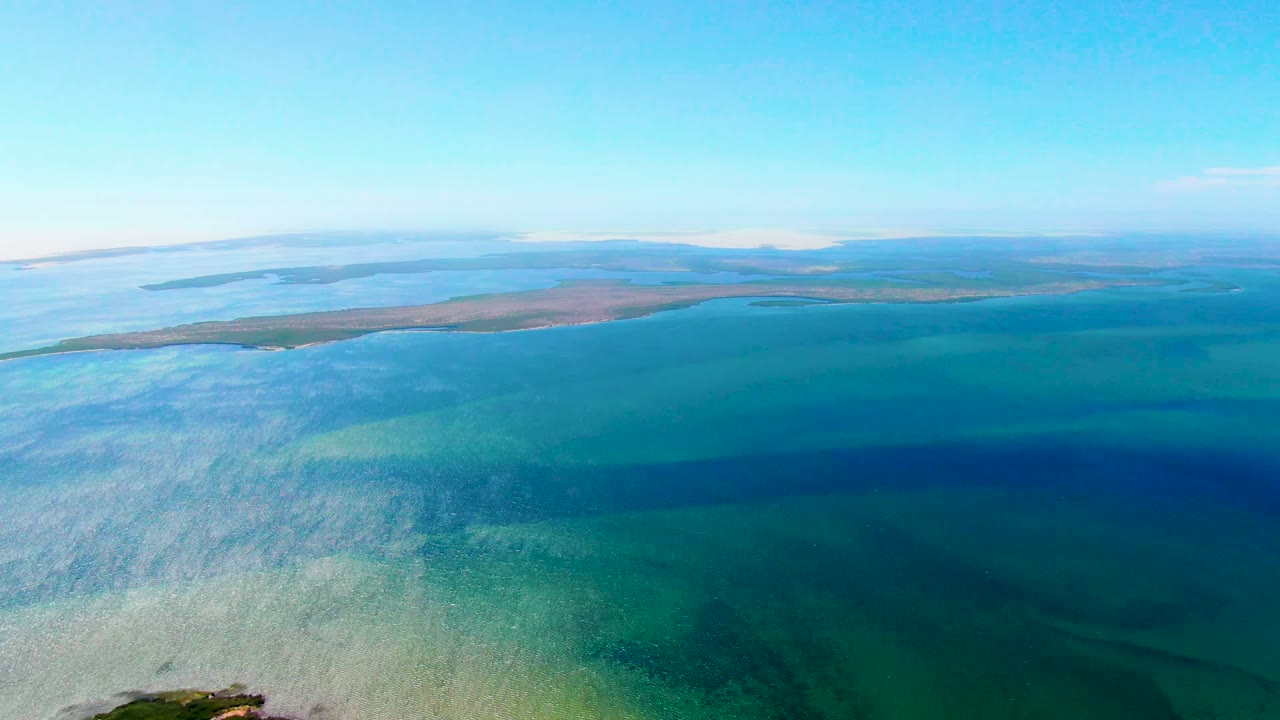 Opening aerial frame over San Carlos, Baja California Sur, with pale turquoise shallows in front, darker blue water beyond, a low island near the horizon, and no visible people.