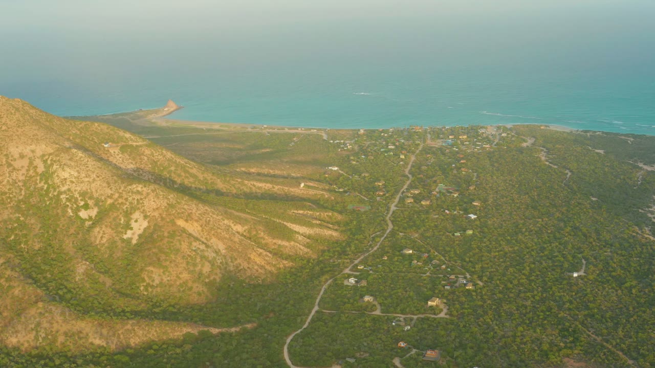 Aerial frame over Cabo Pulmo, Baja California Sur showing a tan mountain slope at left, winding valley roads, scattered buildings, a sandy shoreline, turquoise ocean, and no visible people.