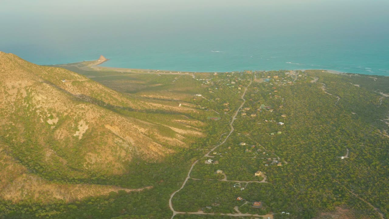 Aerial frame over Cabo Pulmo with the mountain face filling the left foreground, a green developed valley and winding road in the center, turquoise ocean beyond, and no visible people.