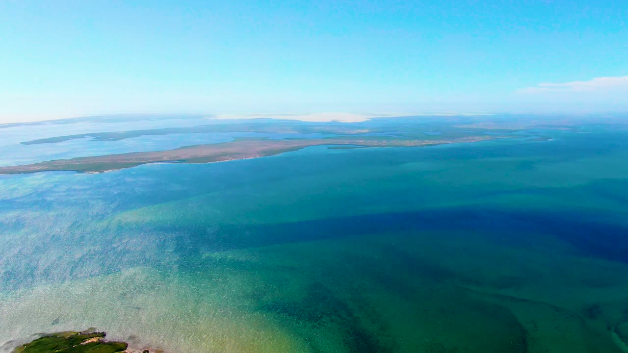 Opening aerial frame over San Carlos, Baja California Sur, with pale shallow water at left, deeper blue-green water in the center, a low sandbar on the horizon, and no visible people.