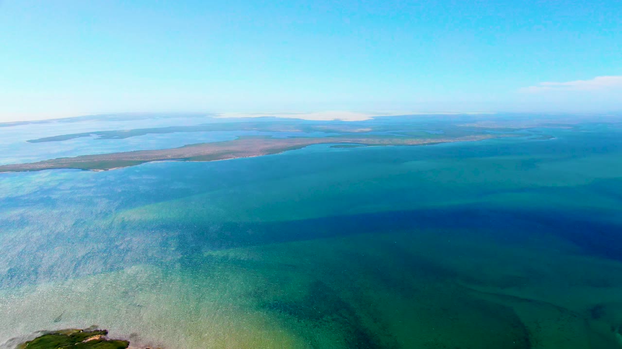 Blue-green ocean shallows with rippled pale water and a low sandbar near the horizon.