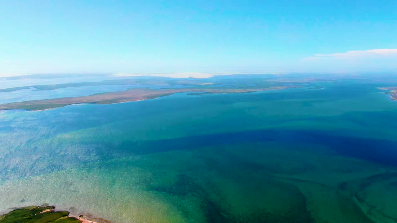 At 0:00, an aerial view over San Carlos, Baja California Sur shows clear turquoise shallows, darker offshore water, distant low land, a small cloud, and no visible people.