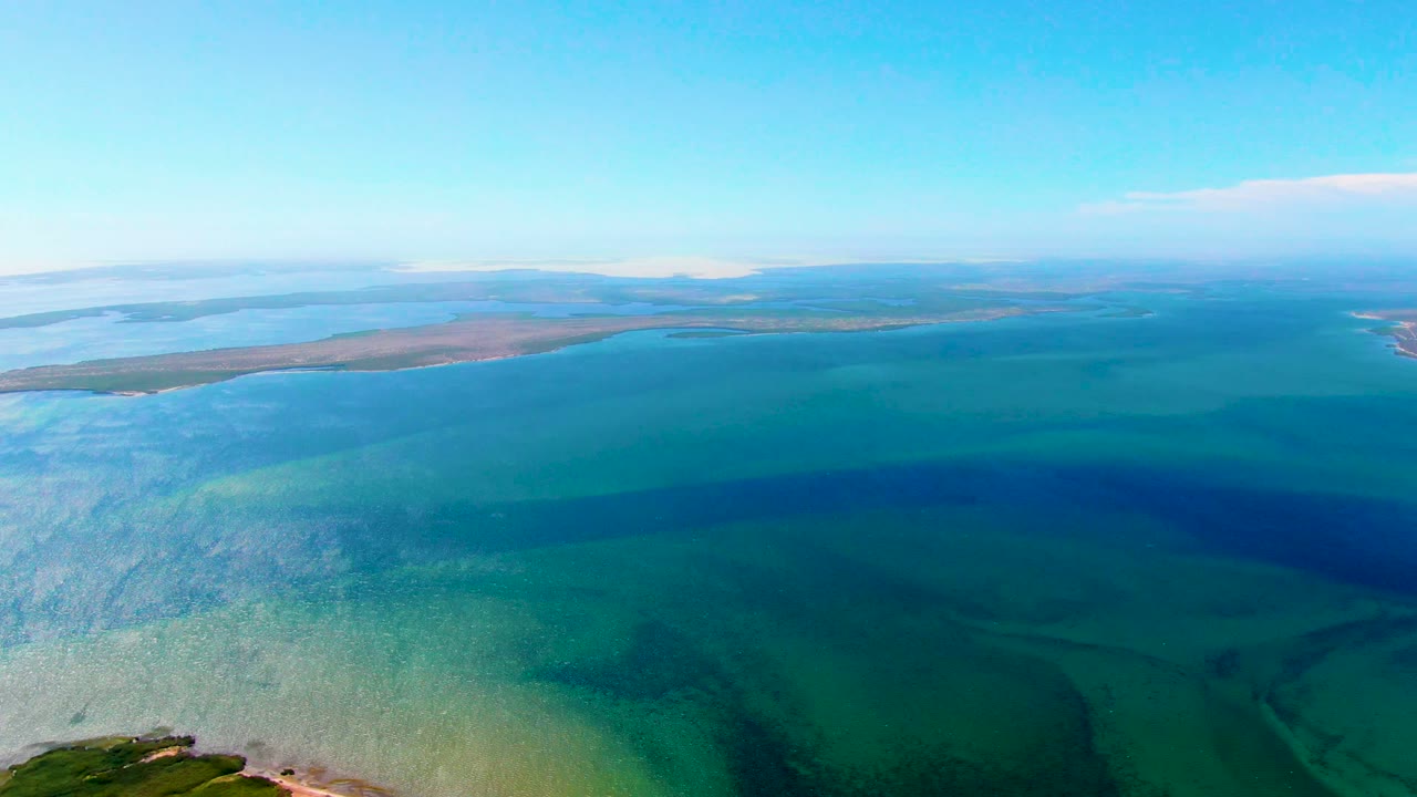 Clear coastal water darkens toward the horizon with low land in the distance.