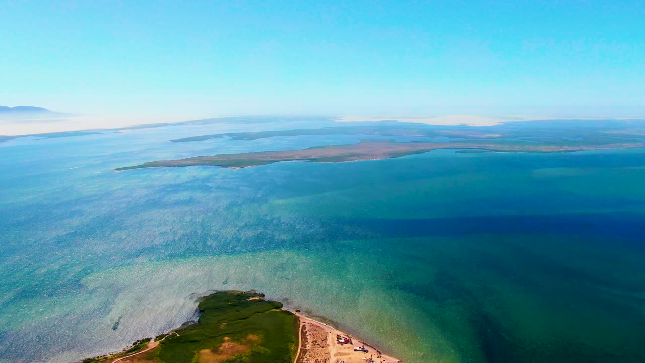 Opening aerial frame over San Carlos, Baja California Sur, with the green peninsula low in frame, turquoise water spreading outward, a distant island on the horizon, and no visible people.
