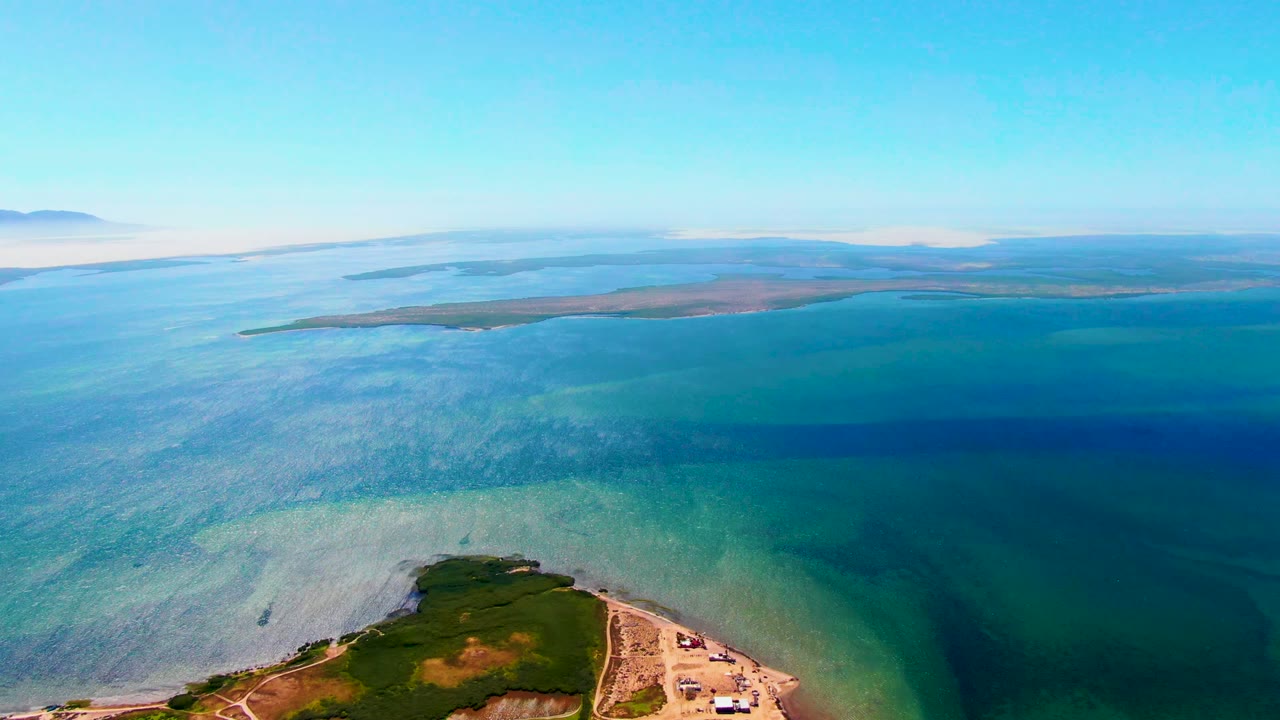 Opening aerial frame over San Carlos, Baja California Sur coastal water, with a green shoreline point at the bottom edge, distant land across the horizon, and no visible people.