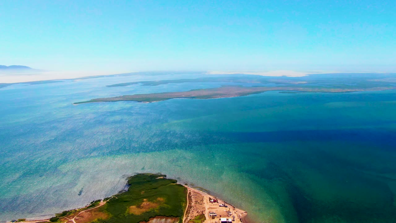 Wide coastal aerial with turquoise shallows, dark blue water, and a distant horizontal landmass.