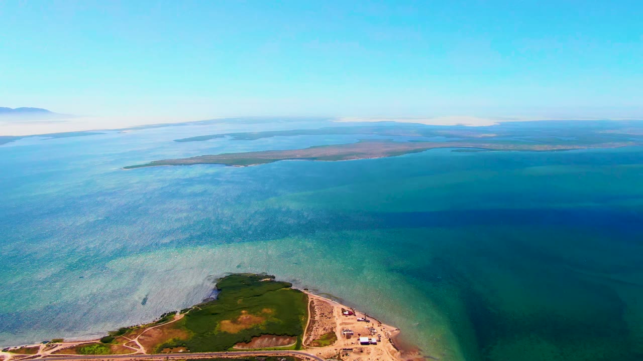 Aerial frame from San Carlos, Baja California Sur, showing a green shoreline point at the bottom, clear turquoise water shifting to deeper blue, distant low land on the horizon, and no visible people.