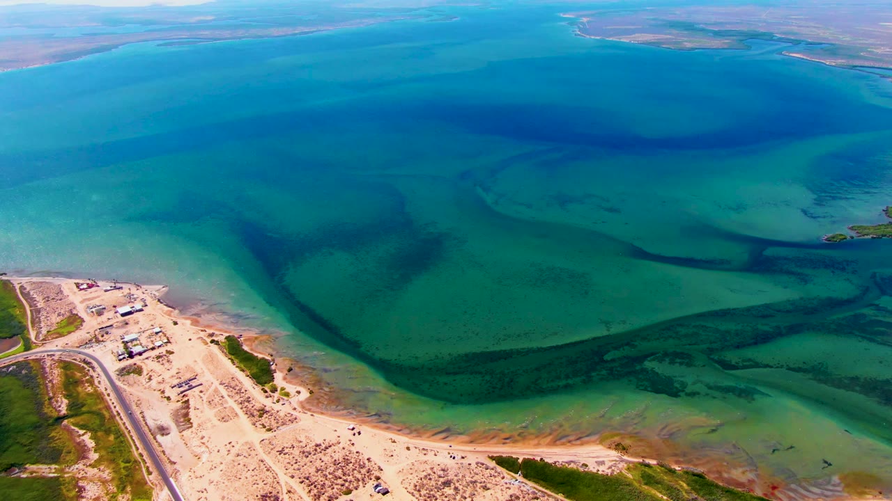 High aerial view over San Carlos, Baja California Sur, with turquoise water, dark current bands, a sandy point, a road along the shore, sparse vegetation, and no visible people.