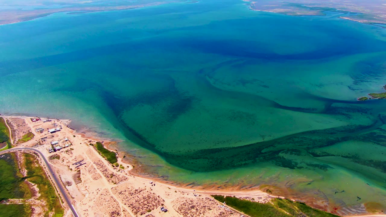 Drone view of shallow green-blue water currents beside a pale beach and road.