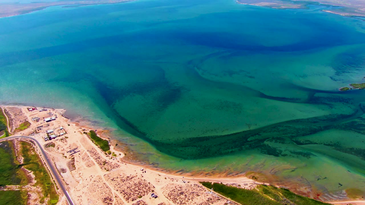 San Carlos aerial view with deep blue water at the top of frame, turquoise shallows through the center, a sandy beach point at lower left, a road, beach vegetation, and no visible people.
