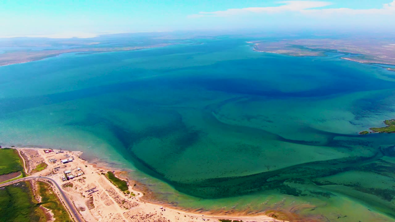 Aerial frame over San Carlos, Baja California Sur, showing blue-green reservoir water, sandy shore, a road, small structures, low vegetation, distant land, and no visible people.