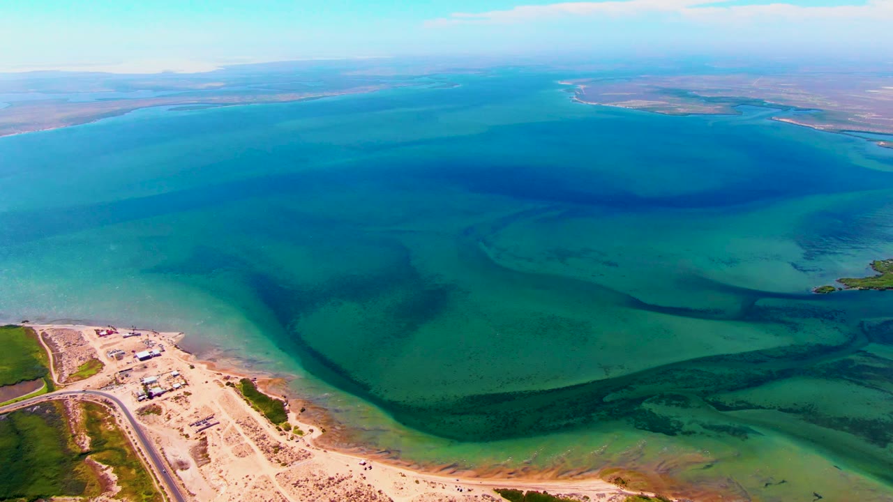 Aerial shoreline view with turquoise shallows, darker water bands, sand, road, and small buildings.