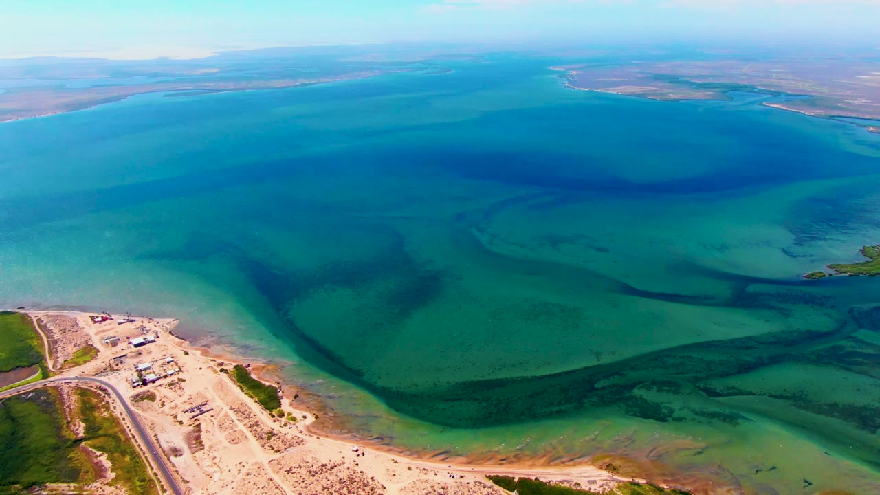A wide San Carlos reservoir frame shows pale sand and a road along the bottom edge, green-blue shallow water, deep blue water farther out, distant landforms, and no visible people.