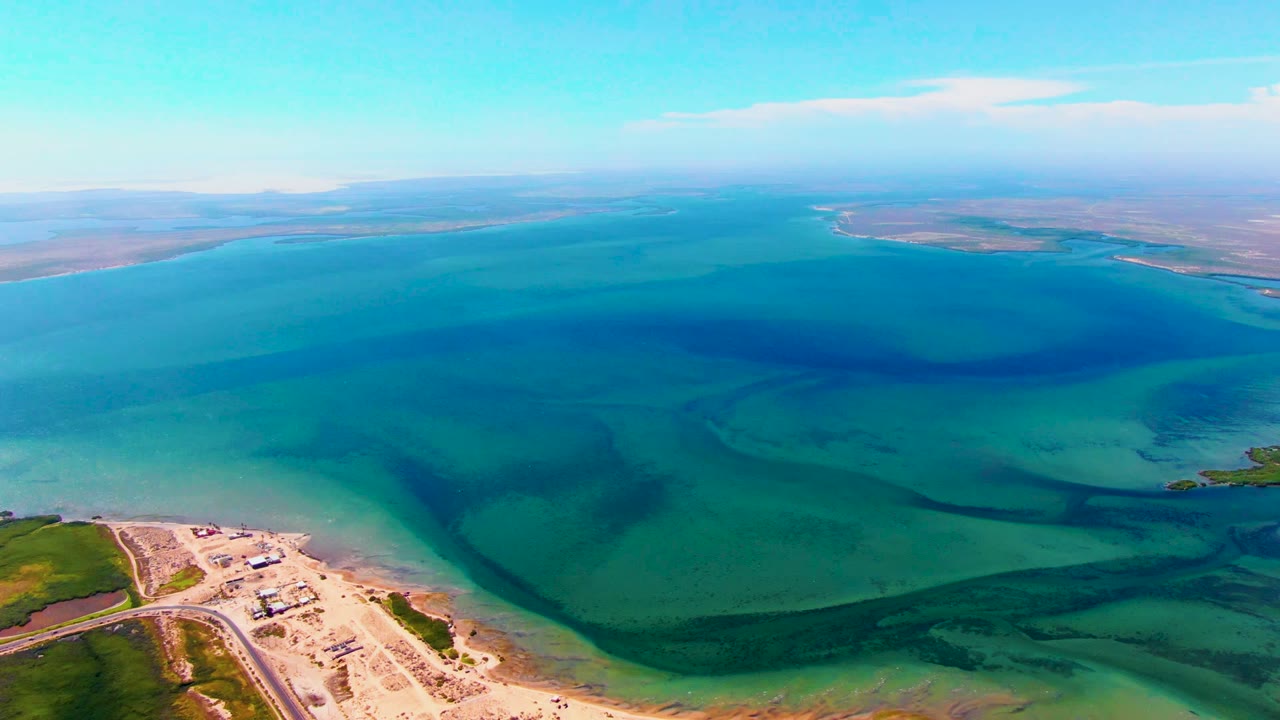 Wide drone frame over San Carlos, Baja California Sur, showing turquoise shallows, darker algae bands, a small sandy shoreline area at lower left, a low horizon, and no visible people.