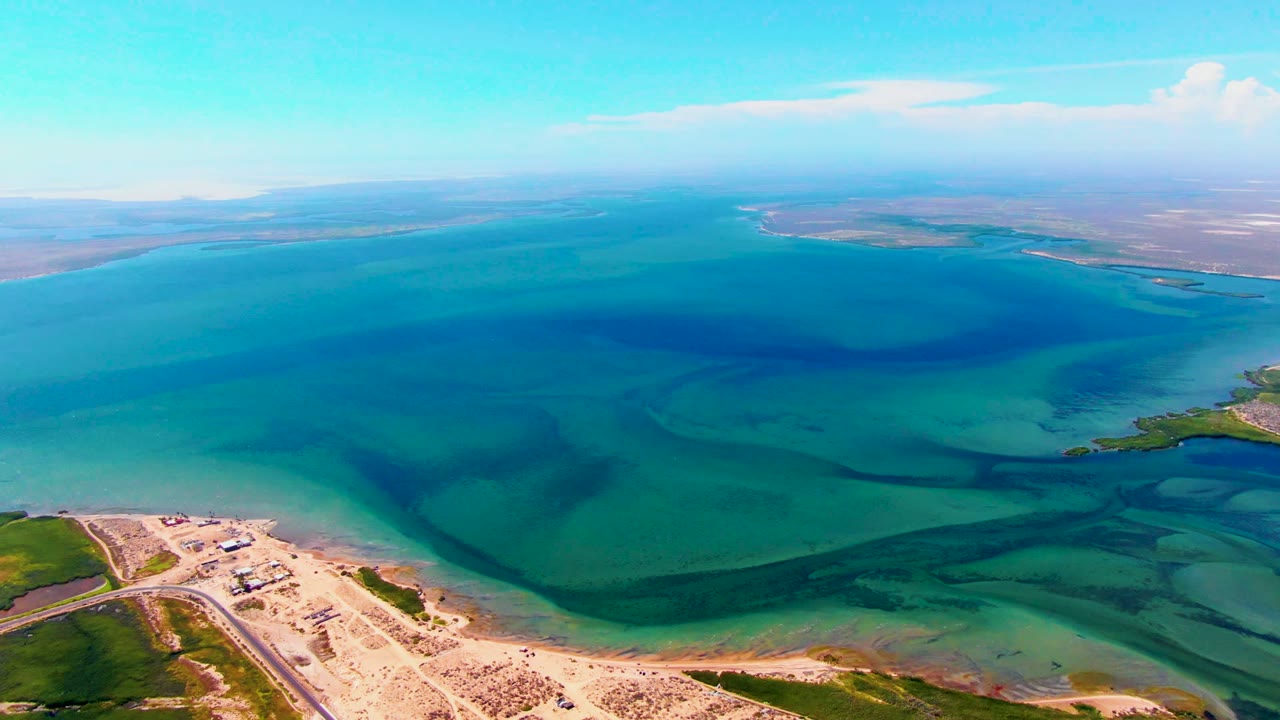 Opening aerial frame over San Carlos, Baja California Sur, showing blue-green water bands, a sandy shoreline corner, green coastal ground, distant land, clear sky, and no visible people.