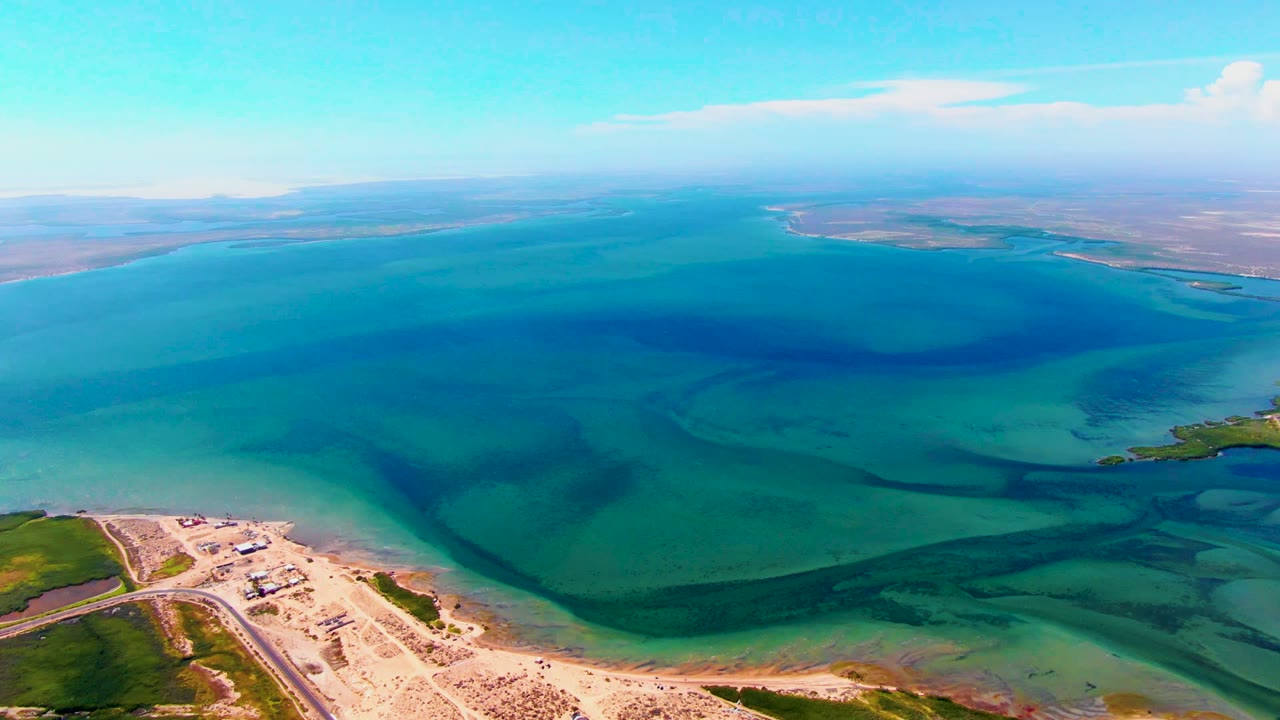 Aerial frame over San Carlos, Baja California Sur, showing the beach and road near the lower left, green shoreline at lower right, wide blue-green water, hazy distant land, and no visible people.
