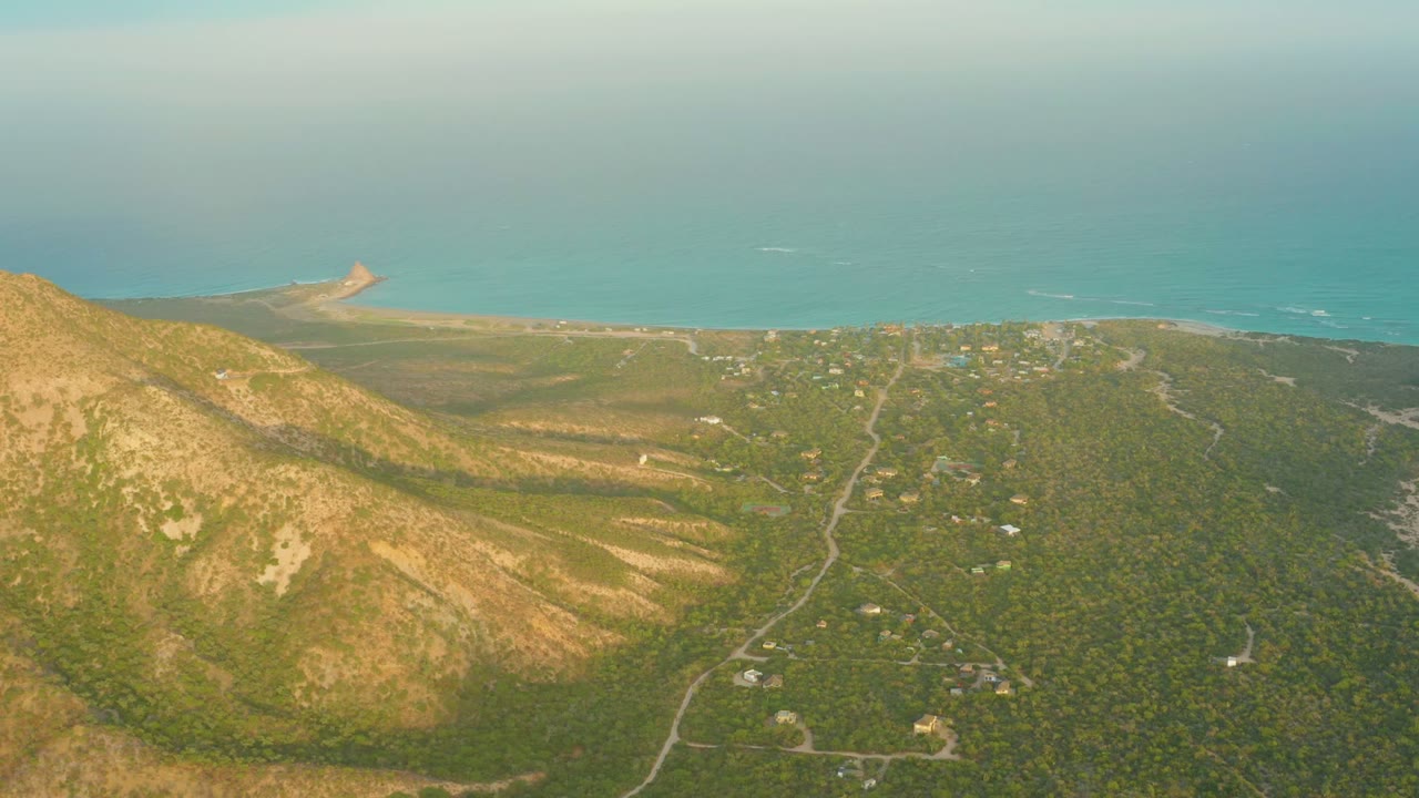 Opening aerial frame over Cabo Pulmo with an ocher mountain ridge at left, green lowland roads below, sandy shore beyond, blue ocean at the horizon, and no visible people.